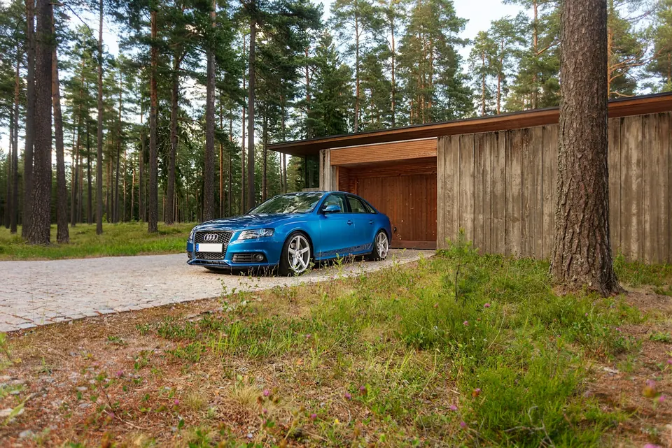 Blå personbil parkert på en brostein-oppkjørsel foran et trehus i en furuskog